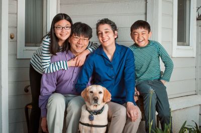 Family portrait outside of a home. A multiracial family. An Asian Man sitting next to a multiracial woman. The left of the man is a young multiracial girl with long hair and glasses. To the right of the woman is a young multiracial boy with short hair. A golden retriever service animal is sitting front and center between the two adults.