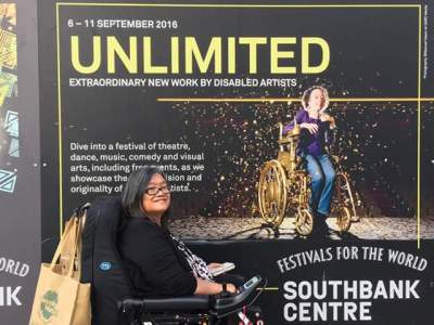 Profile view of an Asian woman in black wheelchair. She has a brown shopping bag hanging on the back of her wheelchair. Behind her is a large billboard for "Assisted Suicide: The Musical" showing at the Southbank Centre in London. The word UNLIMITED is in gold. The billboard shows a thin petite white woman in a gold manual wheelchair sprinkled in gold confetti.