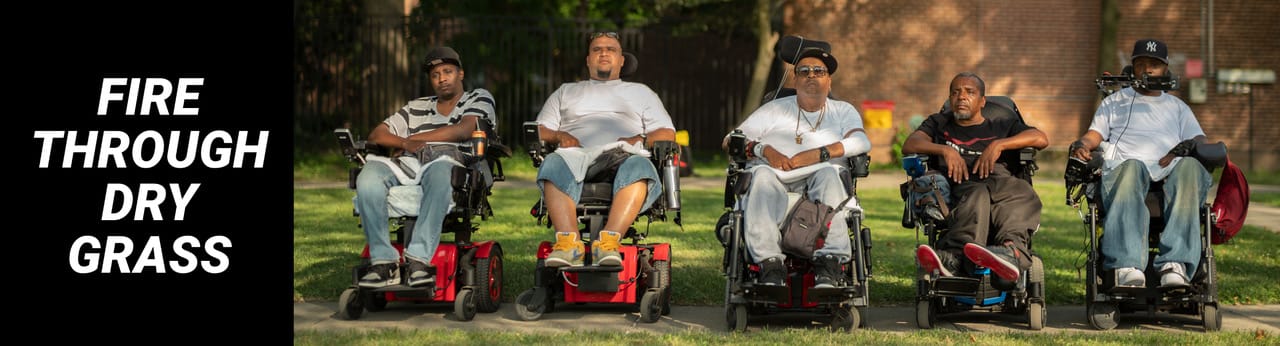 Five Black and brown men (from left to right: Vince, Jay, Tito, Pete and Var) sit in wheelchairs outside, all lined up in a straight horizontal line facing the camera. They wear T-shirts and sneakers and some wear baseball caps. They all look into the camera with serious expressions. Photo credit: Elias Williams