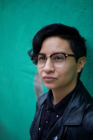 Image of a lightskinned Indigenous and white queer with short black hair, black-rimmed glasses, a button-up black polo, and a black pleather jacket leaning against a green wall and looking away from the camera. Image by Neha Gautam Photography.