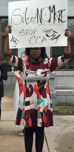 A Black woman wearing glasses wearing a multicolored dress holding a white cardboard sign that reads "Silent No More."