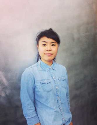 Portrait photo of an Asian American woman looking directly in the camera with a slight half smile. She is wearing a light blue button-up shirt and a long earring on her right ear. She has a lip ring, dark eyes, and dark hair that is tied up with bangs that are cut in a diagonal line, slanting to her left. She is set against a silver background.