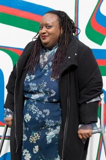 Imani stands on her crutches in a blue dress and jacket in front of a blue red and green mural wall. Photo credit: Madasyn Andrews
