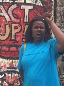 A Black woman with her left fist in the air in a sign of solidarity and Black power. She is wearing a teal colored t-shirt.