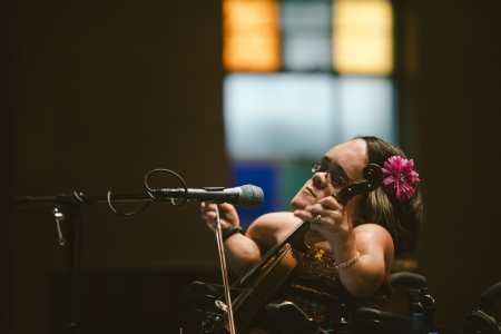 Caucasian woman of small stature with dark brown hair. She is playing a violin positioned in front of her like a cello. Her eyes are closed, deep in concentration. Photo credit: Mark Brown