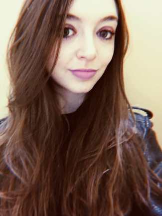 A light-skinned woman with long wavy brown hair stands, smiling. Her face fills almost the entire photo frame. She has brown-colored eyes, brown eyeshadow, and purple lipstick while wearing a black leather jacket. Behind her is an off-white colored wall.