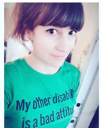 Pictured is a Mestiza woman with big brown eyes and pale skin color. She is positioned at a side angle with respect to the camera and is wearing a green t-shirt that reads "My other disability is a bad attitude". 