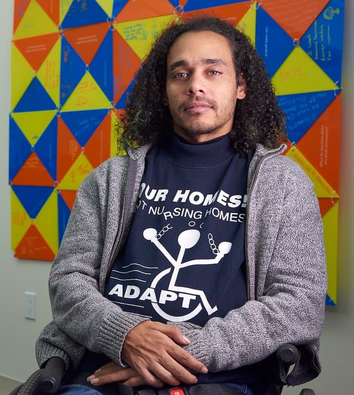 Full front photo of Germán Parodi. A Puerto Rican man sitting in his wheelchair with a slight grin and hands crossed on his lap. He has brown skin, and shoulder length curly dark brown hair. He is wearing a black long sleeve shirt that says “Our Homes Not Nursing Homes ADAPT ” on it and a gray zip-up sweater on top. Behind him appears a painting with multi-colored triangles