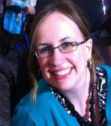 A white woman with glasses and long brown hair smiling at the camera. She is eating a turquoise jacket and a black necklace.