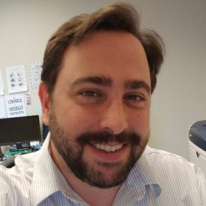 Image Description: A smiling white disabled man with brown hair and a beard wearing a white and grey striped dress shirt. on the office walls behind him are several signs, the only readable one saying 'The Struggle is Socially Constructed'.