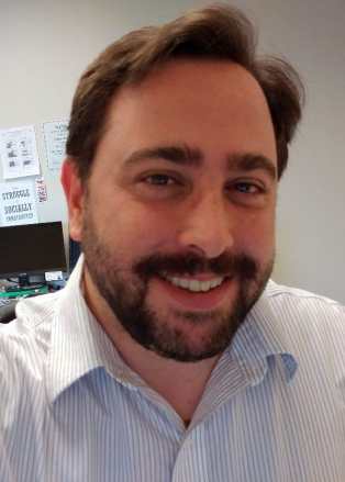 Gregg Beratan, a white man with brown hair and a brown beard. He is smiling and wearing a white shirt with narrow gray stripes.