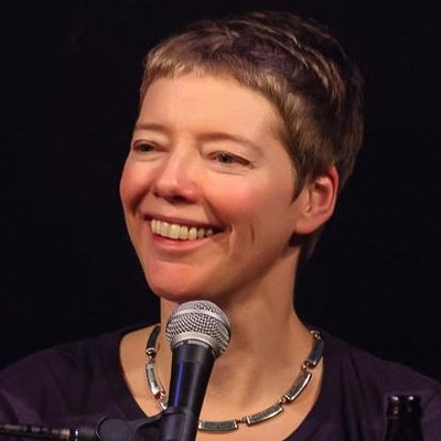 A middle-aged white woman with short cropped hair. She is looking toward the left of the image with a microphone in front of her. The background is all black and she is wearing a scoop neck shirt with a metallic necklace.