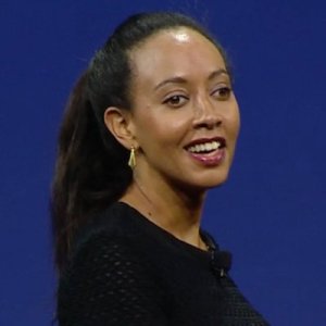 Photo with a blue background with a young Black woman with long hair pulled back. Her body is turned at an angle toward the camera. She is wearing a black sweater, gold earrings. She is smiling.