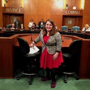 Image description: Maria Town, a young white woman with long brown hair standing behind a room that looks like a hearing room in the city hall of Houston, TX. She is wearing a bright red dress with a black and white blazer.