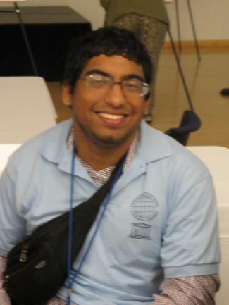 Hamza Jaka, a brown American Pakistani man, smiles up at the camera. He is wearing glasses, a blue UNESCO Chair Institute shirt, and a fanny pack along with lanyard.