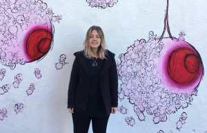 A young Caucasian woman with short blonde/pink hair is standing and smiling, wearing a camouflage jacket, in front of a white wall outside with a mural painted on it. The mural consists of large pink and red breasts hanging from vines, made out of fetuses.