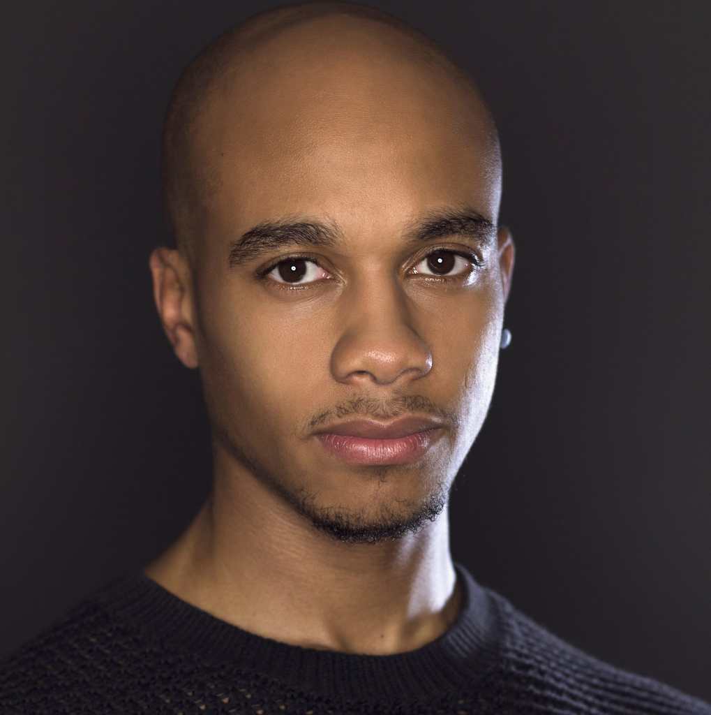 Hari Ziyad, a Black person with a bald head and short facial hair, stands in front of a dark grey background looking into the camera with a slight smile. They are shown from the shoulders up wearing a black knitted sweater, with a black circular earring in their left ear. Photo by Brandon Nick