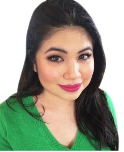  A picture of a woman sitting. Image is cut off at the chest. She is looking straight at the camera, and smiling with bright pink lipstick and pink blush on her cheeks. Long black hair is asymmetrical and falls to the right side of her face. She has long lashes and brown eyes. Wearing a soft, green sweater. Background of picture is white--no furniture.