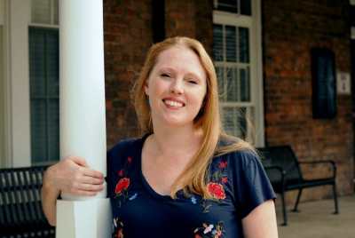 A white woman with long, straight blondish-reddish hair. She smiles at the camera, eyes slightly squinted in the sunlight, with one arm wrapped around a narrow pillar on a brick building. Her shirt is gray with a flower pattern. There are benches on the porch behind her.