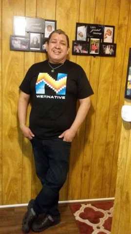 Picture of Hector standing in the hallway of his house. The walls and floors are wooden and there are some pictures of his family on the wall. He is standing and is wearing a T-shirt that reads WeRNative that has an native patterns. He is wearing dark blue jeans and great Crocks. His hands are in his pant pockets and he is wearing a lime green bracelet that reads “Each Mind Matters”. He is smiling at the camera and is wearing a turquoise and silver pendant on a small chain around his neck.