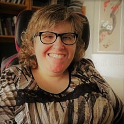 A photo of Heidi, a white woman wearing black-rimmed glasses and smiling brightly. She is surrounded by books and art.
