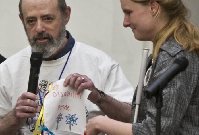 Photo of an older white man who is bald with a beard holding a microphone. He is holding a drawing that has flowers and the words 'Disability Pride.' Next to him is a young woman with long blond hair and a white cane. She is also holding a drawing up.