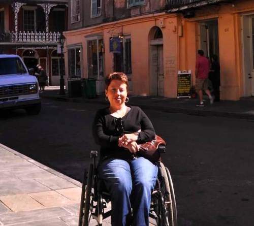 Outdoor photo of a white woman in the French Quarter of New Orleans, Louisiana. She has short, cropped brown hair, gold earrings and is wearing a long-sleeved black shirt and blue denim jeans. She is sitting in a manual chair and there are historic buildings behind her. 