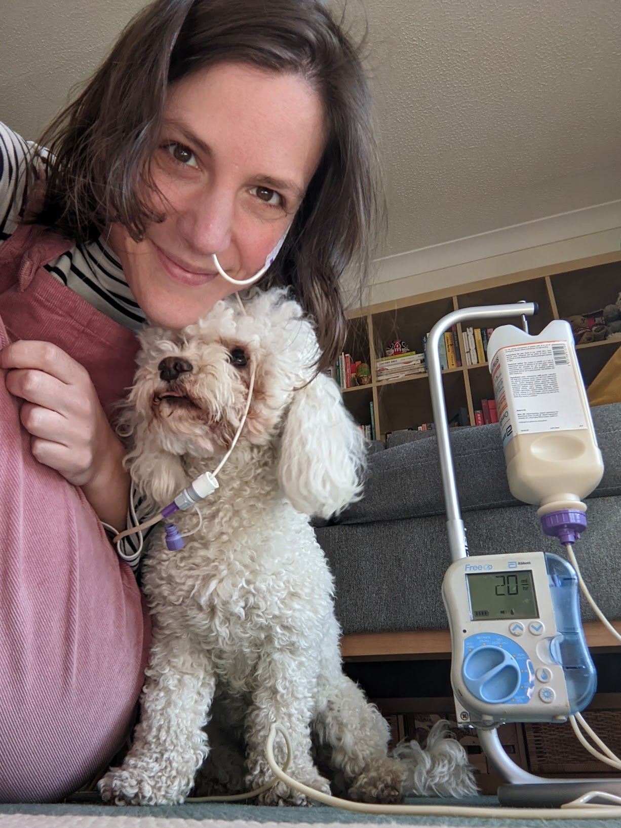 Background with a light pink background. Selfie of a white woman with dark hair and eyes with an NJ tube on her face, next to a white toy poodle, and with her feed hanging on a frame with a pump attached.
