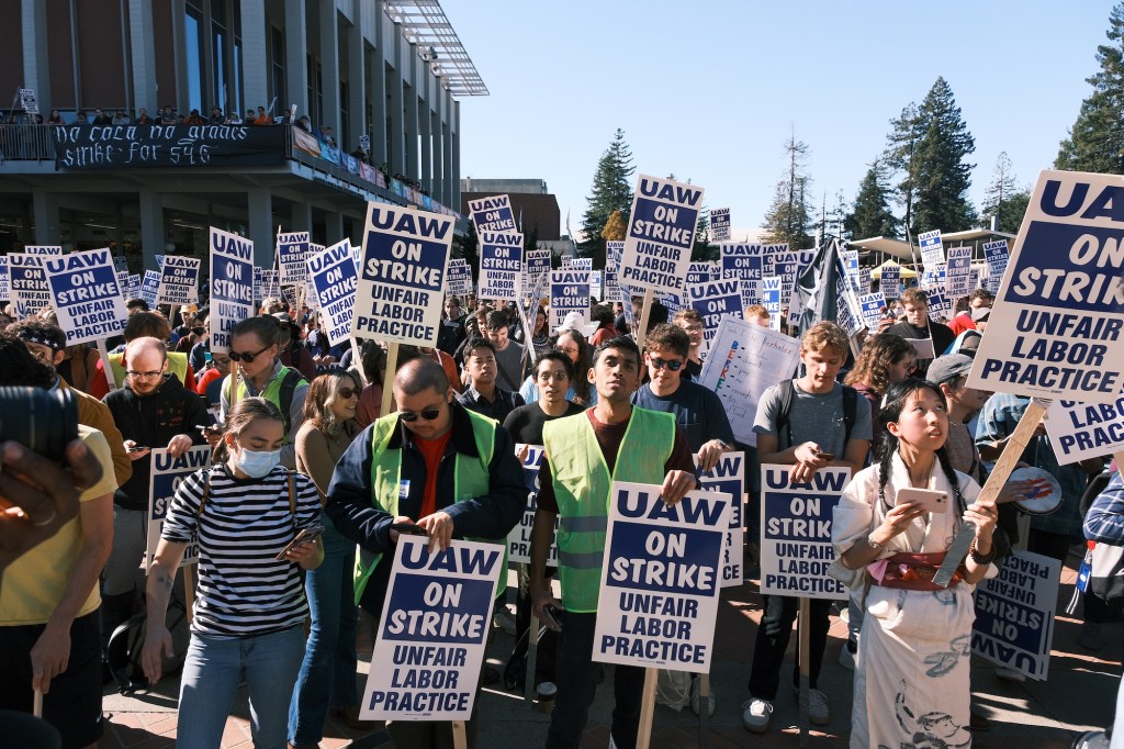Many, many workers with picket signs reading "UAW: On Strike: Unfair Labor Practice". They are crowded together outdoors and in the sea of faces there appears to be only one worker wearing a surgical mask. Photo by: Ian Castro. 2022. Provided on FairUC Now Website for Press Use