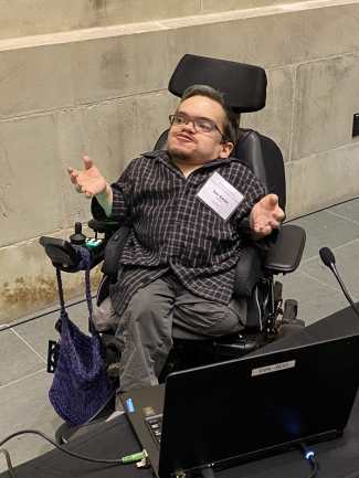 Ian is a white man in his 30s sitting in a power wheelchair with oene leg crossed under the other. He's spreading his hands and grinning, one eyebrow slightly raised, like he's gesturing mid-explanation. Nearby him is a laptop and a table with microphones.