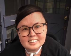 A mixed race Korean and white queer person smiles head on at the camera. She has big glasses and is wearing a crewneck. Her trach and wheelchair can be seen.