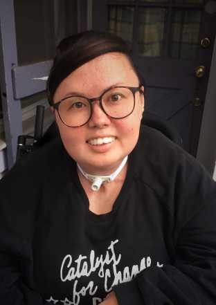 A mixed race Korean and white queer person smiles head on at the camera. She has big glasses and is wearing a crewneck. Her trach and wheelchair can be seen.