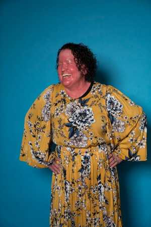 Image shows Carly standing, hands on hips, laughing in front of a blue background wearing an awesome bright yellow dress adorned by beautiful white flowers.