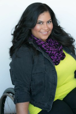 Image description: Danielle Perez sits in her wheelchair. Her long black hair is blown out. Danielle is smiling and wearing a neon green top, with a purple and black stripped scarf, and a black denim jacket.