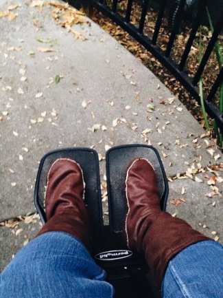 A sidewalk with bits of leaves on the ground. In the foreground, a pair of legs with denim jeans wearing soft brown leather boot socks. The feet are resting on a wheelchair's footrests.