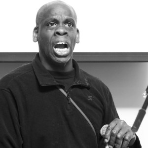 Black and white image of a middle-aged Black disabled man wearing a dark shirt, with a cane nearby his hand.