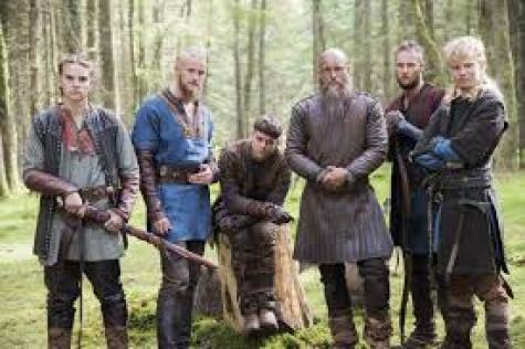 Image description: screenshot from an episode of Vikings, an original series on the History Channel. Ivar the Boneless is a young Viking warrior with short dark hair. He is in the center, sitting on a tree stump. To the left of him are his brothers Hvitserk and Bjorn. To his right is his father Ragnar and brothers Ubbe and Sigurd. 
