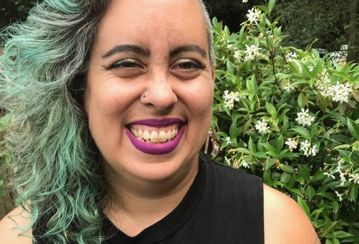 Leah looks at the camera in a garden in South Seattle in August. She has long curly brown, silver and green hair, dark magenta lipstick and light brown skin, and is grinning in front of a garden wall covered in blooming jasmine. Photo credit: Jesse Manuel Graves