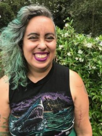 Leah looks at the camera in a garden in South Seattle in August. She has long curly brown, silver and green hair, dark magenta lipstick and light brown skin, and is grinning in front of a garden wall covered in blooming jasmine. Photo credit: Jesse Manuel Graves