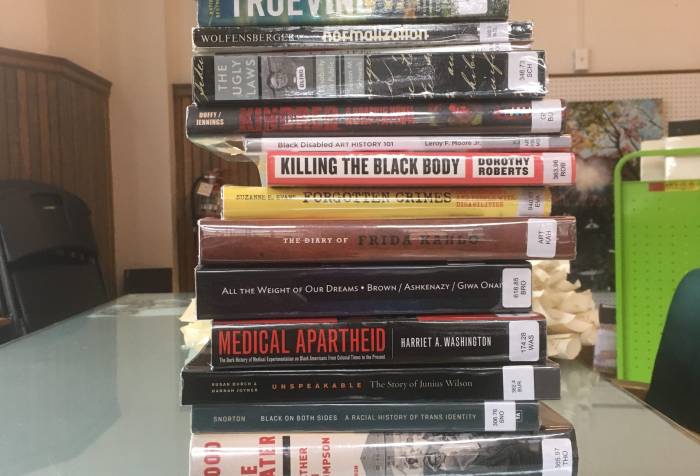 A stack of 17 library books lying horizontally. Text on the spines of the books from top to bottom: The Girl with All the Gifts, Disability and Difference in Global Contexts, Truevine, Normalization, The Ugly Laws, Kindred, Black Disabled Art History 101, Killing the Black Body, Forgotten Crimes, The Diary of Frida Kahlo, All the Weight of Our Dreams, Medical Apartheid, Unspeakable: The Story of Junnius Wilson, Black on Both Sides: A Racial History of Trans Identity, Blood in the Water, Sick from Freedom, Thinking Class
