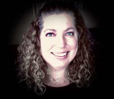 A headshot of an adult woman with curly, brown-blonde, hair, green eyes, and a large smile.