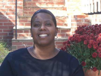 Image description: Woman smiling and sitting on brick stairs in black shirt with flowers behind her. This is Dara Baldwin - African American woman, social justice activist and disability rights advocate.