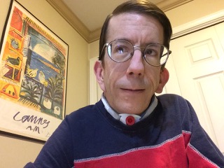 Image description: Portrait photo of a white man, clean shaven, with a tracheostomy, and short brown hair, wearing glasses and a blue and red shirt.