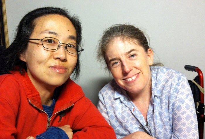 Asian American woman in a wheelchair sitting on the left side. She's wearing a red hoodie and glasses. Next to her is a white middle-aged woman with her hair pulled back and wearing a floral-print shirt. Both are smiling at the camera.