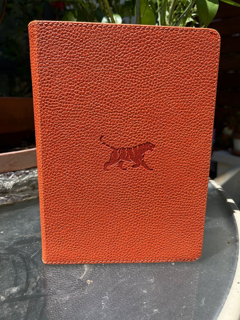 An orange journal with a pebble-like texture on the cover with a tiger in the center. The journal is standing on top of a patio table.
