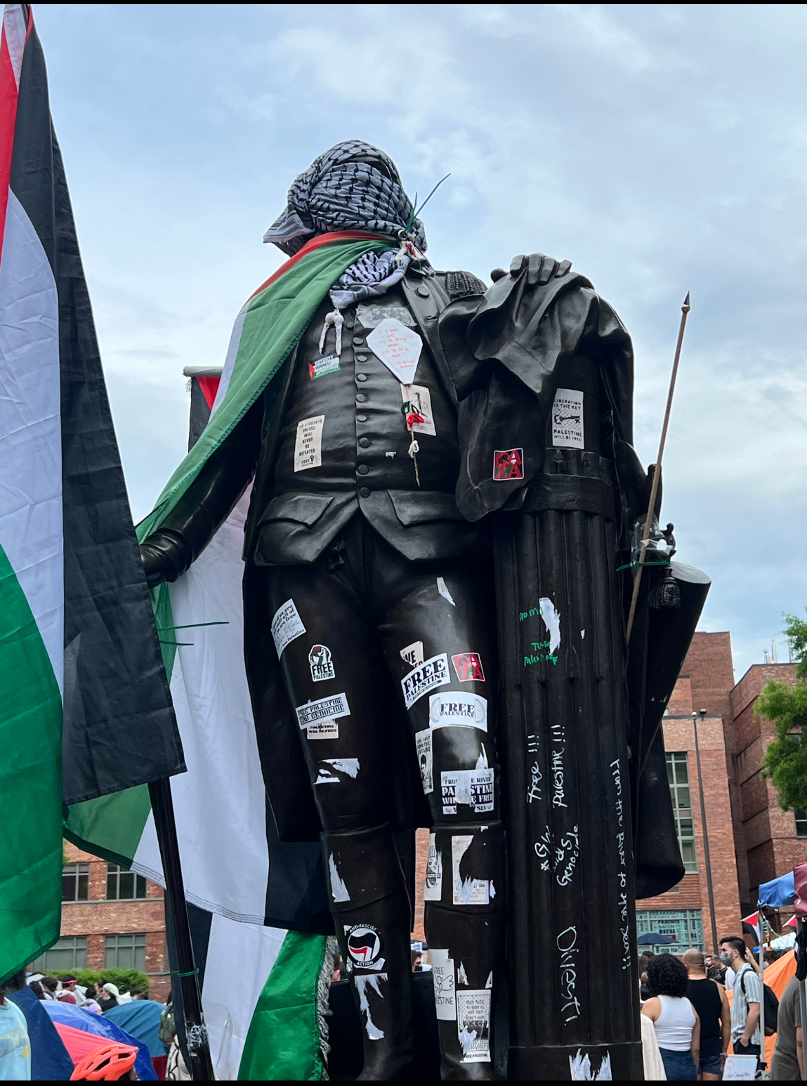 A statue of George Washington in Washington, DC. The statue's face is wrapped in a kuffiyah, it is wearing a Palestinian flag as a cape, and is holding another Palestinian flag. There are various stickers on the chest and body.