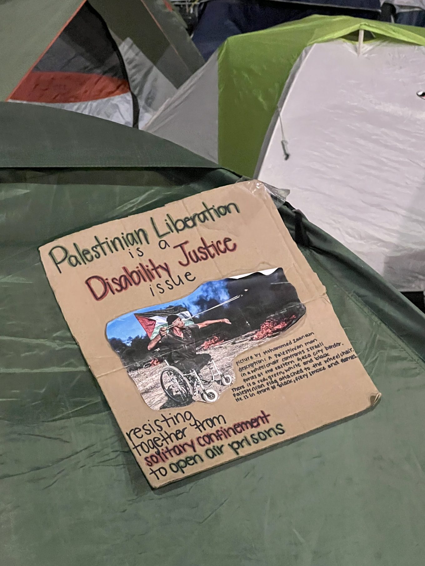 A cardboard sign on a tent at a student encampment in DC. It says at the top in red and black letters "Palestinian Liberation is a Disability Justice issue."  In the center is a photo with the image description: "Picture by Mohammed Zaanoun" Description: A Palestinian man in a wheelchair confronts Israeli forces at the eastern Gaza City border. There is a red, green, white, and black Palestinian flag attached to the wheelchair. He is in front of black, fiery smoke and flames." Below the photo in black, red, and green lettering are the words "Resisting together from solitary confinement to open air prisons."