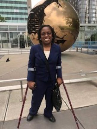 A Black woman in a blue suit standing on crutches in front of a golden replica of the globe outside of the United Nations headquarters in New York.