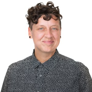 A smiling non-binary white person with short curly brown hair wears black earring plugs and a black button up shirt with small white polka dots