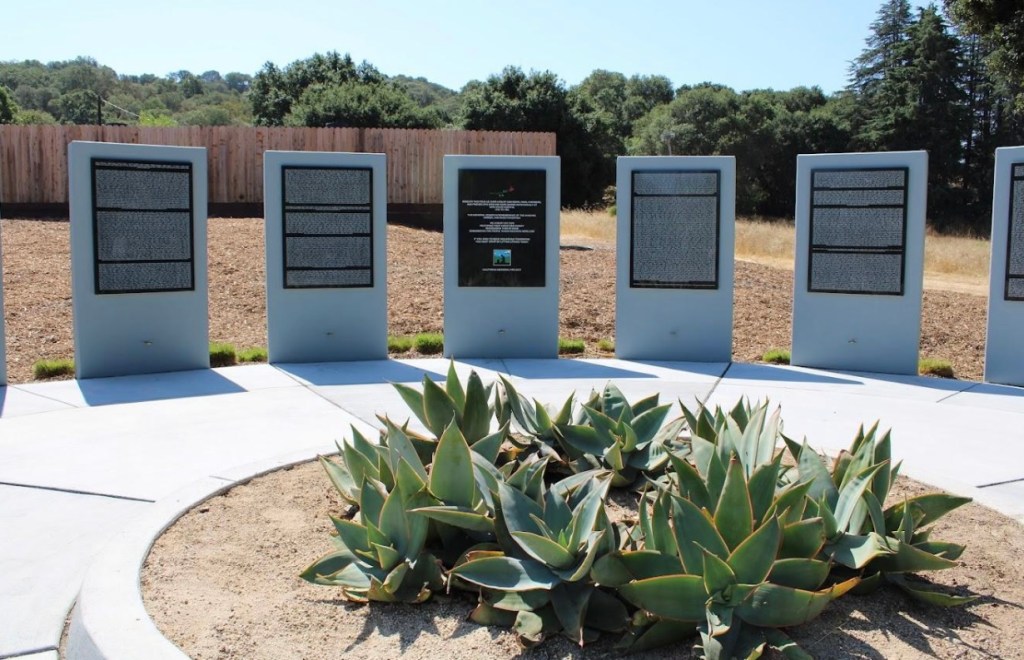 A monument on the grounds of Napa State Hospital in Northern California. It consists of several gray pillars with black displays with white texts, too far away to read. In the foreground are several large spiky succulent plants. In the background are a wooden fence and trees underneath a blue sky.